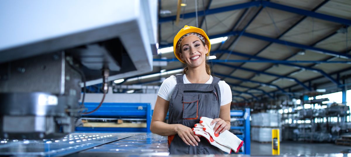 Portrait of female factory worker in protective uniform and hardhat standing by industrial machine at production line. Dia das mulheres - principais desafios